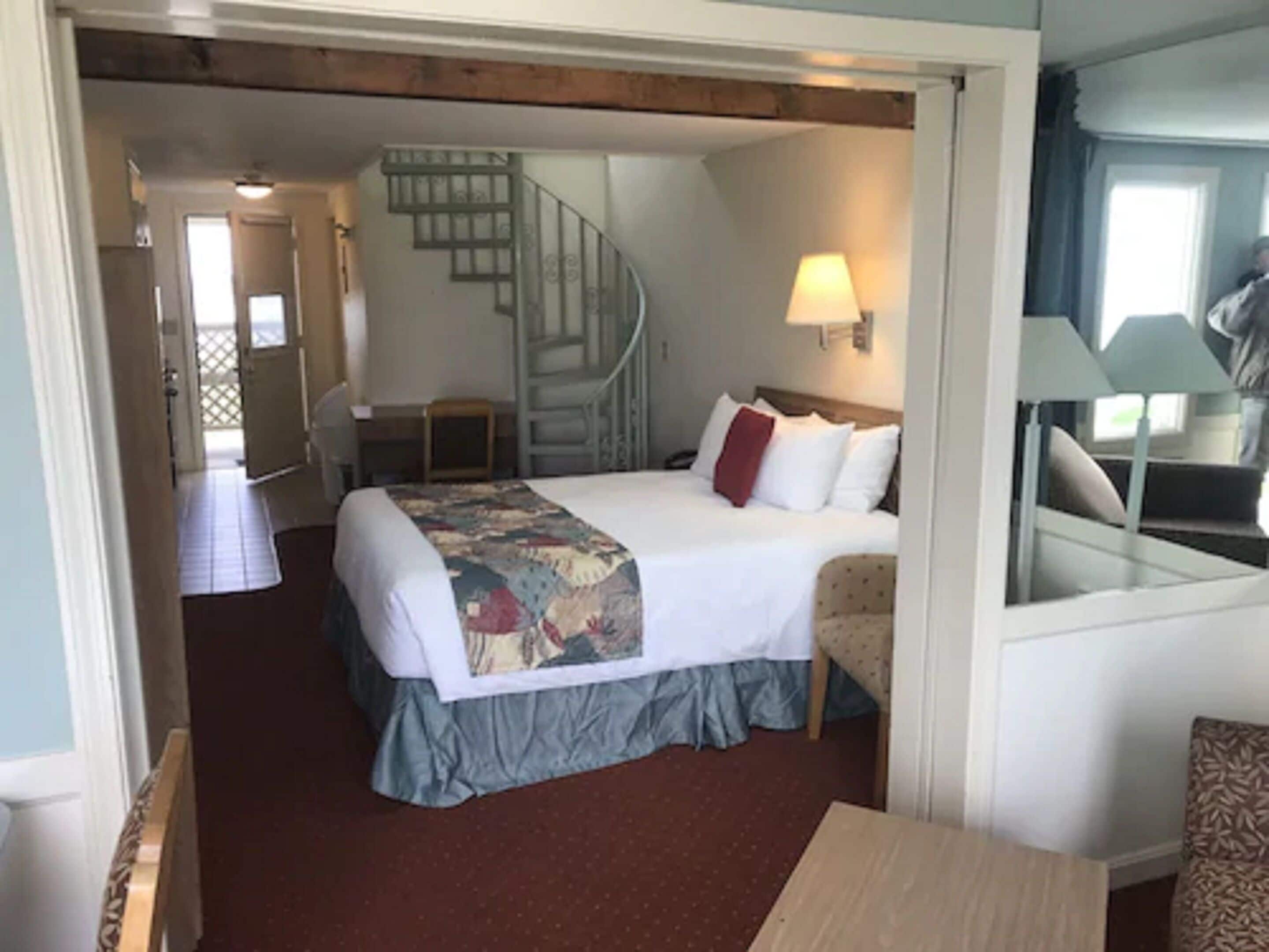 A hotel room with a bed, a chair, a desk, a lamp, and a staircase.