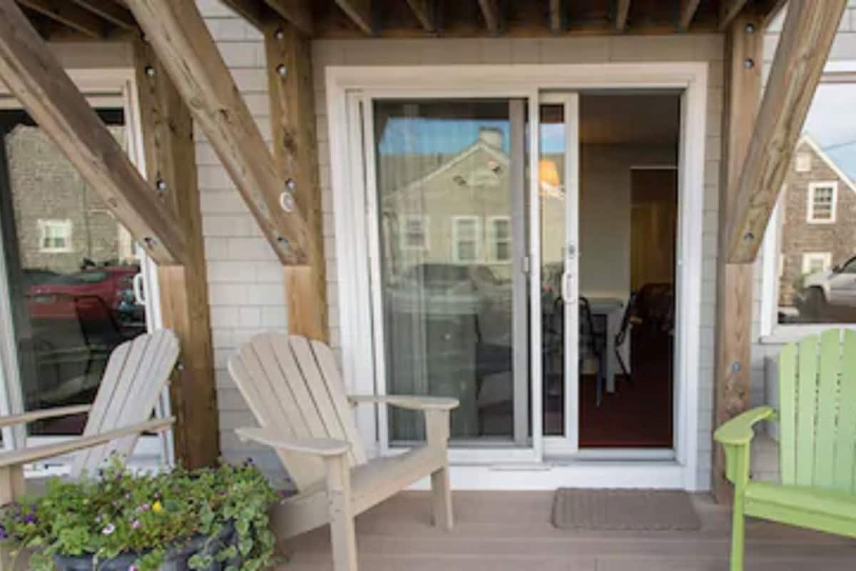 A covered porch with two Adirondack chairs, a sliding glass door, and a brick wall.