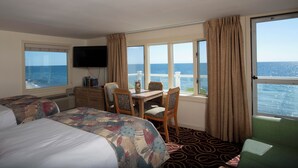 Ein Hotelzimmer mit zwei Betten, einem Esstisch mit Stühlen, einem an der Wand befestigten Fernseher und großen Fenstern mit Meerblick.
