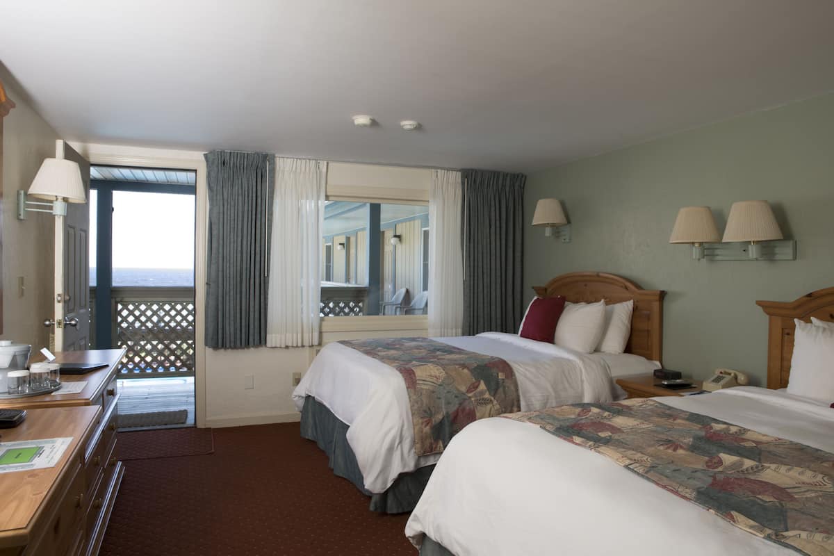 A hotel room with two beds, a desk with a telephone, and a view of the sea through a door.