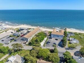 An aerial view of a coastal area with a beach, a parking lot, and buildings. - The Corsair & Cross Rip Oceanfront Resort (Dennis Port)
