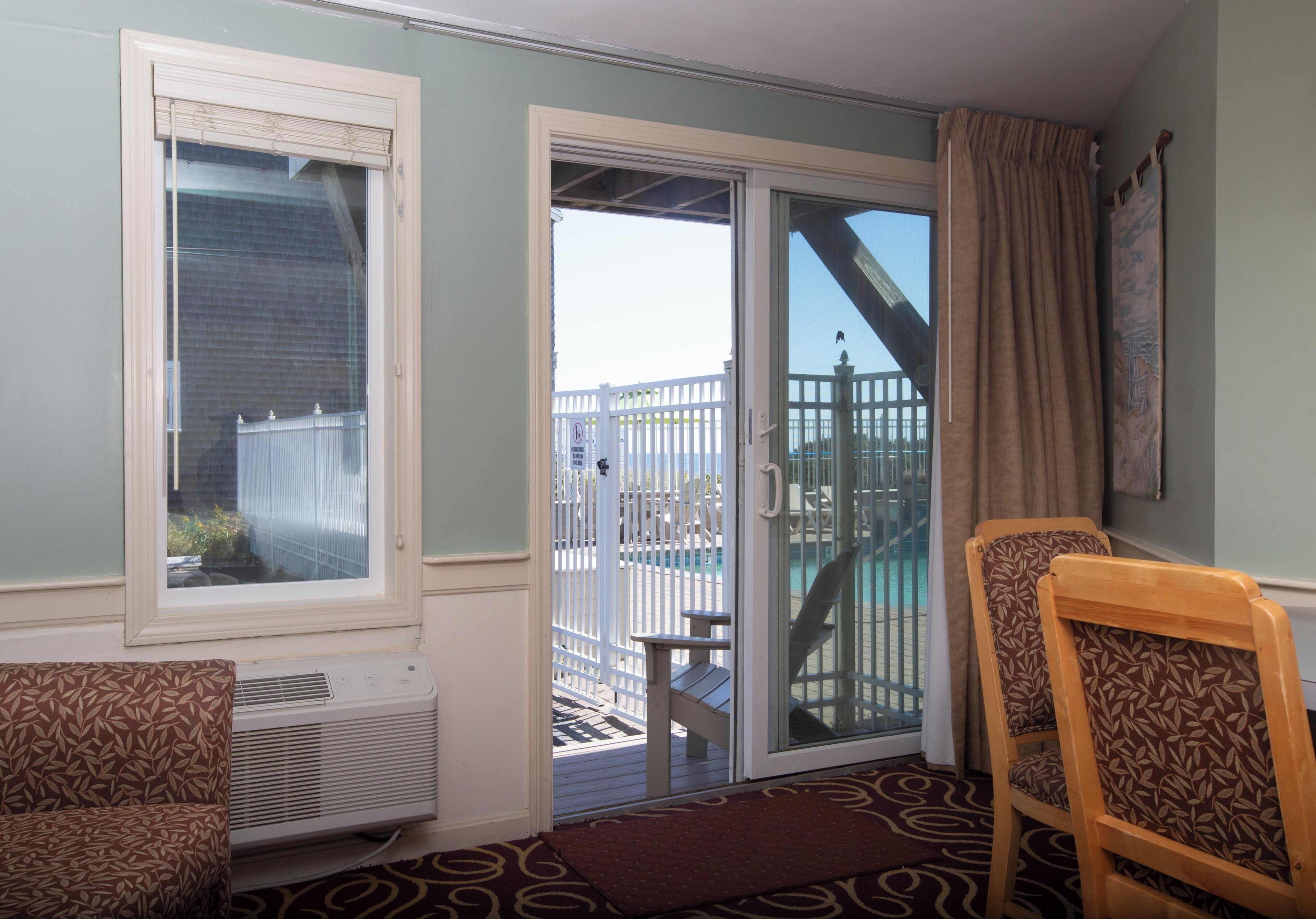 A room with a sliding glass door leading to a balcony, a small table, two patterned chairs, and a wall-mounted air conditioning unit.