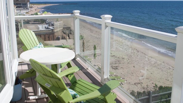 Eine Holzterrasse mit einem Tisch und zwei grünen Stühlen mit Blick auf einen Strand, im Hintergrund ein Zaun und ein Gebäude.