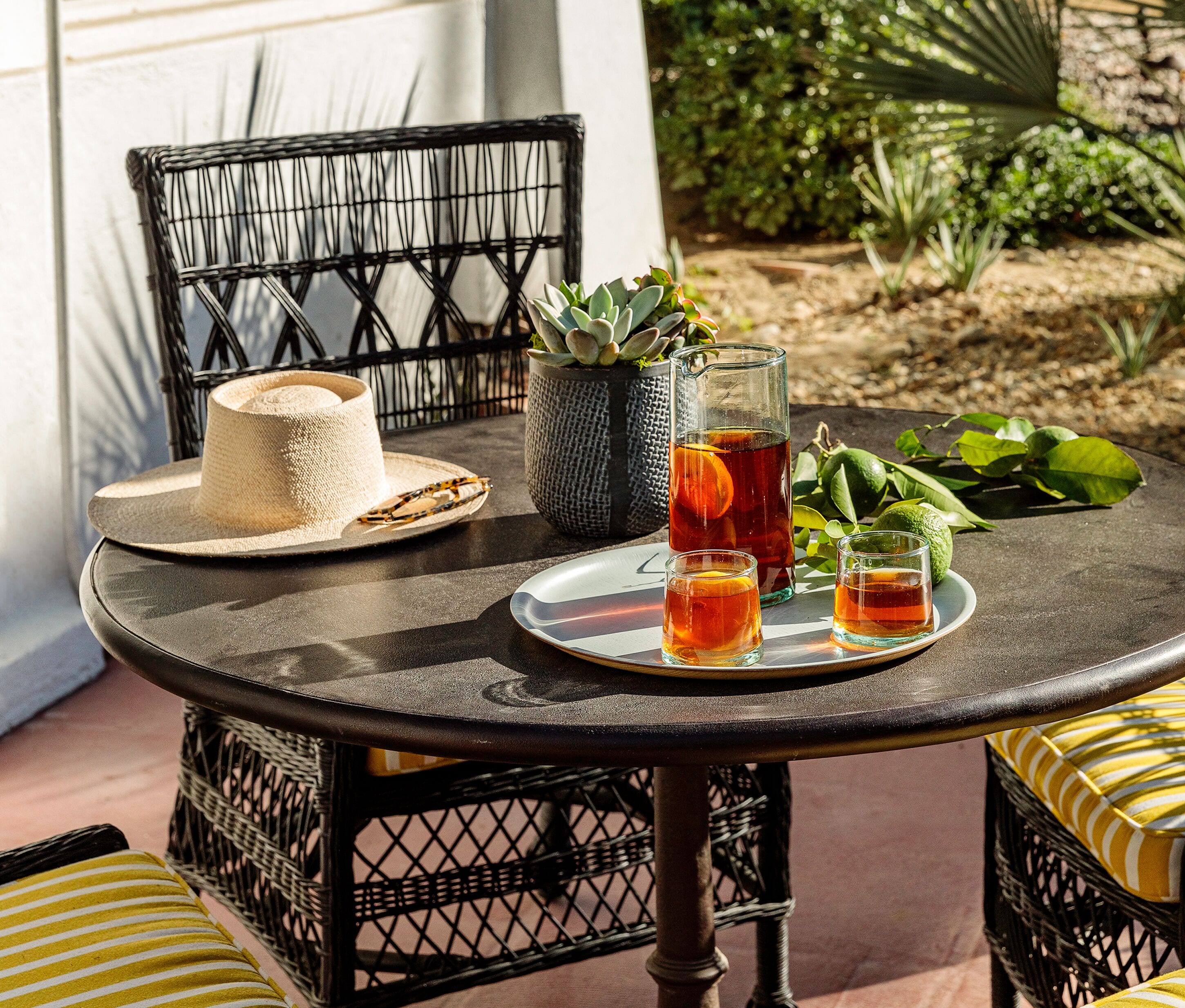 A round table with a pitcher of iced tea, two glasses, a straw, a straw hat, and a woven basket.