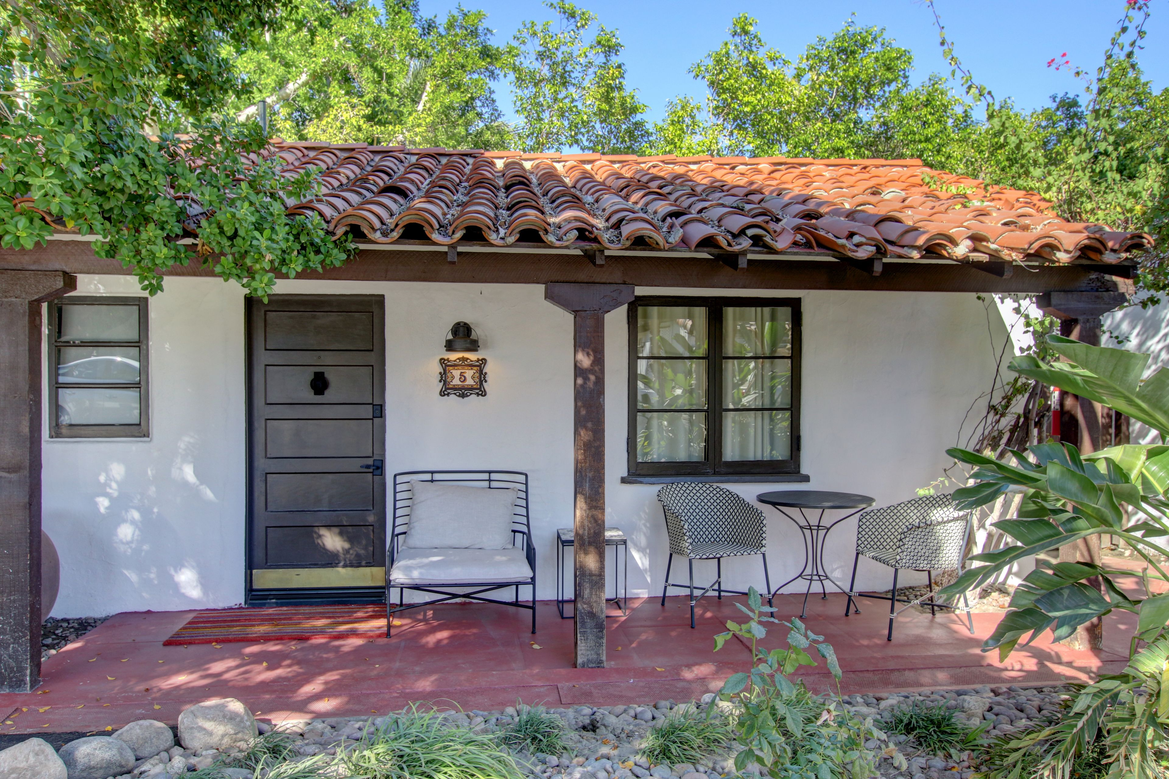 A house with a tiled roof, a door, and two windows. There are two chairs and a small table outside.
