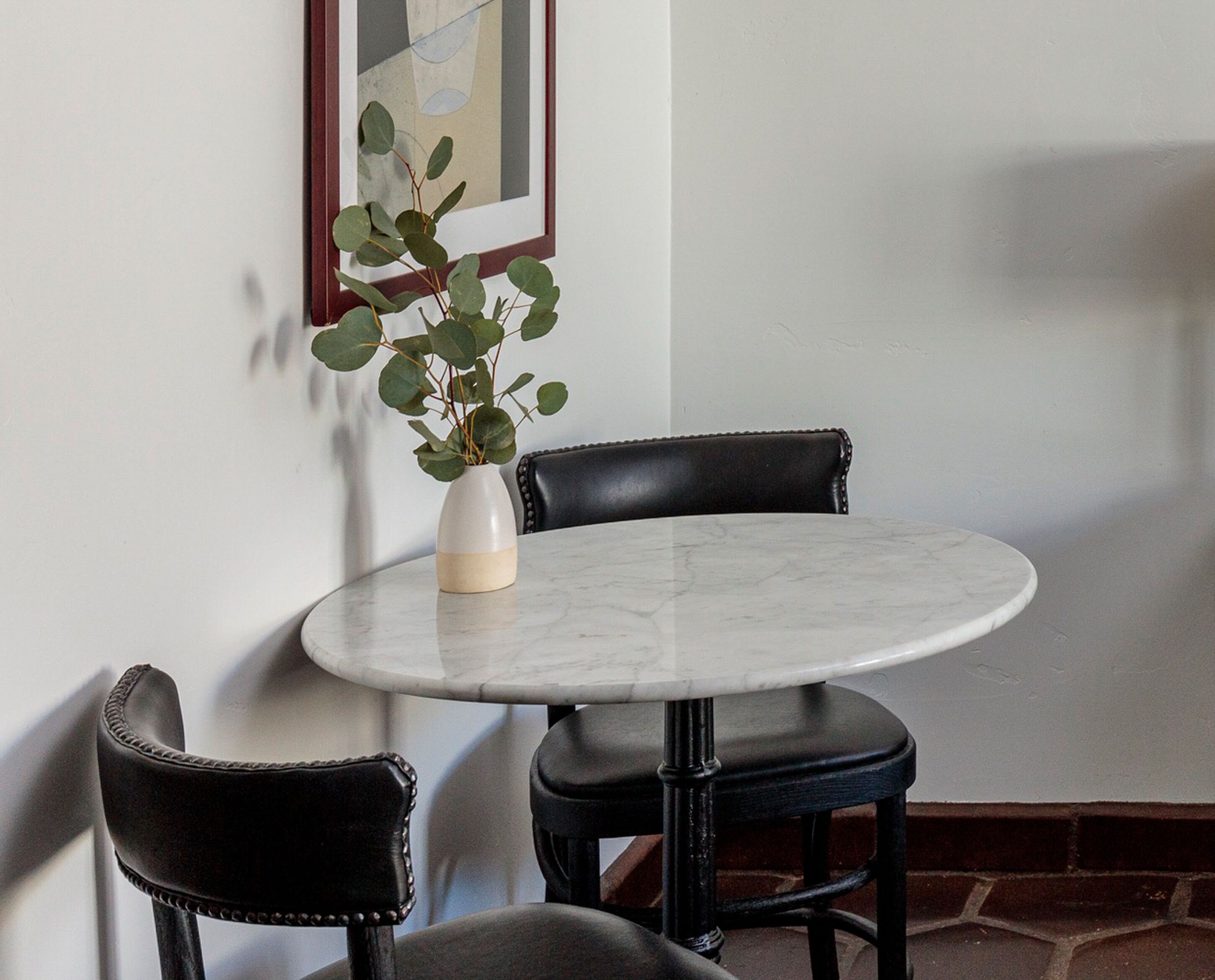 A small round table with a vase of green leaves, two black leather chairs, and a framed picture on the wall.