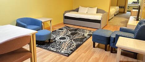 A compact hotel room with a bed, two blue armchairs, a small table, and a patterned rug.