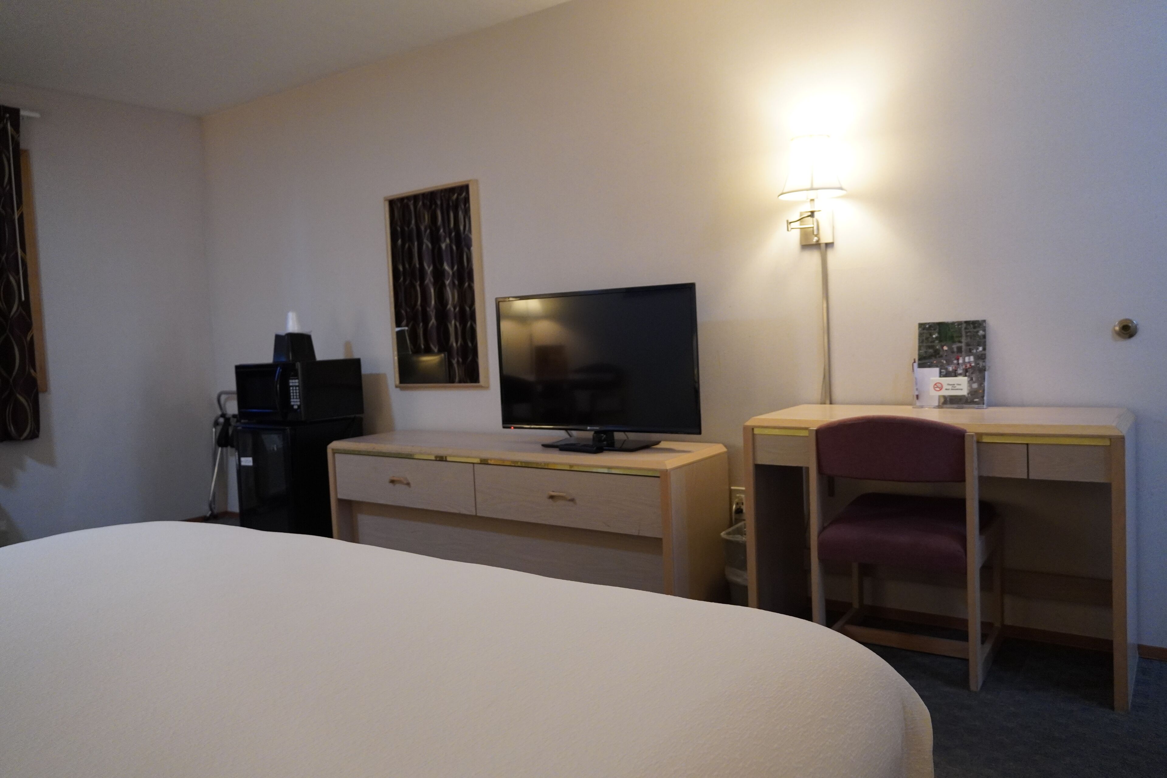 Ett hotellrum med en säng, ett skrivbord med en stol, en tv och en lampa.