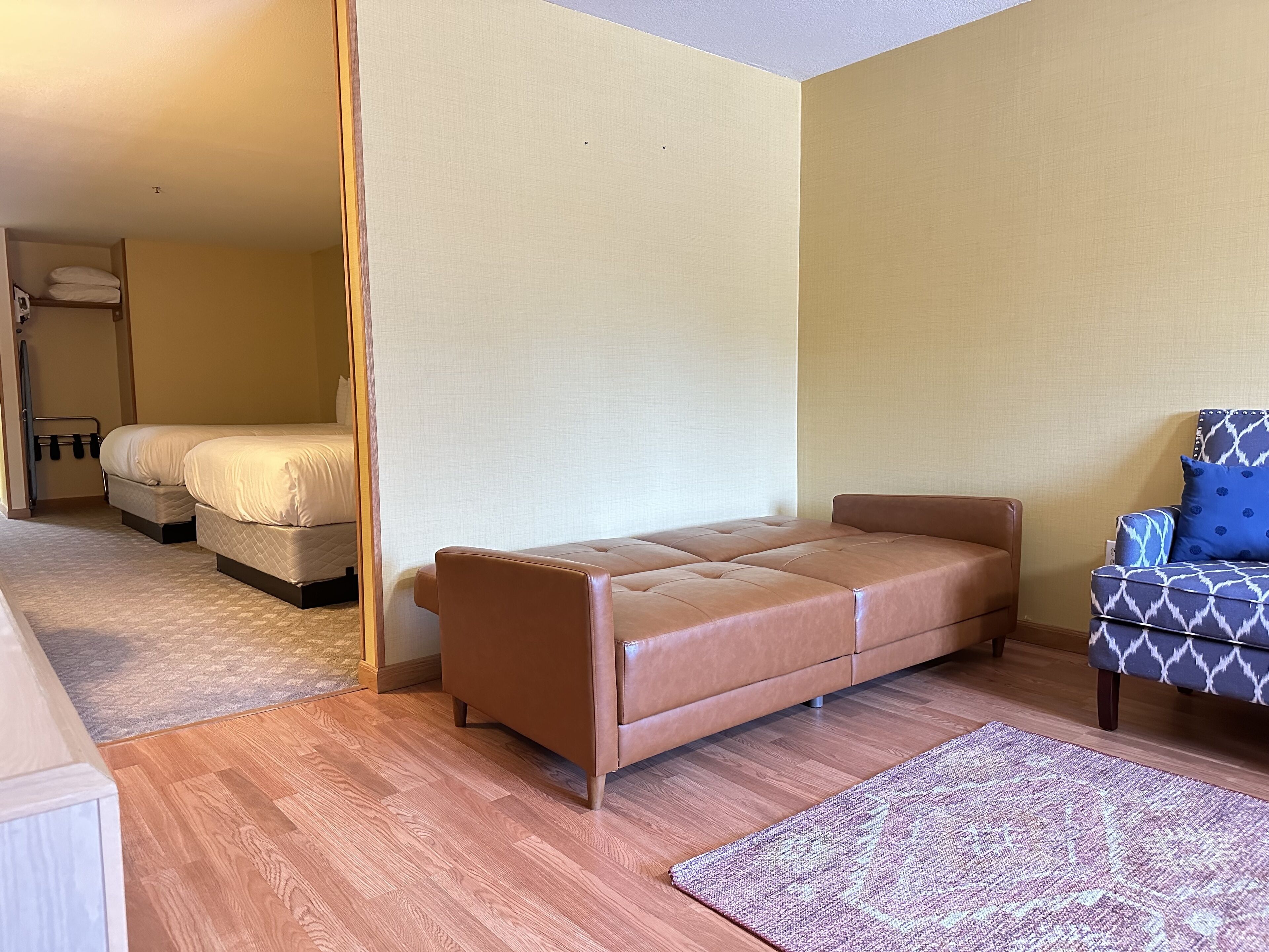 A compact room with a brown leather sofa, a patterned armchair, and a bed with white bedding.
