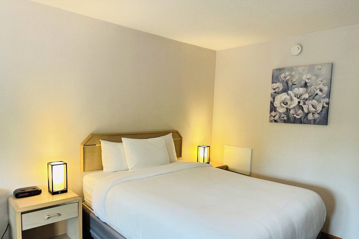 A neatly made bed with white linens, two bedside tables with lamps, a small drawer, and a wall-mounted painting of flowers.