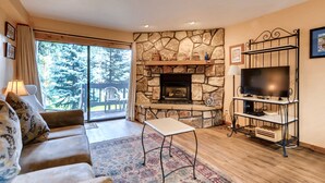 A living room with a stone fireplace, a sofa, a television on a stand, and a view of trees through large windows. - Atrium Condominiums (Breckenridge)