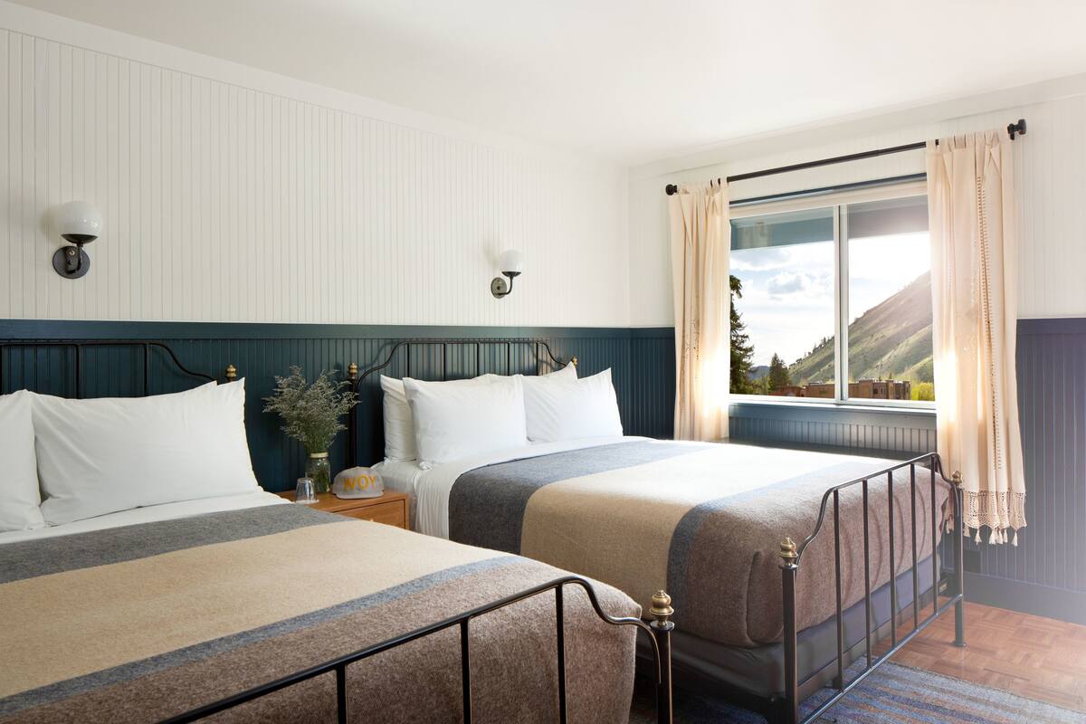 Une chambre d’hôtel avec deux lits, une table de chevet, un vase avec des fleurs et une fenêtre avec des rideaux.