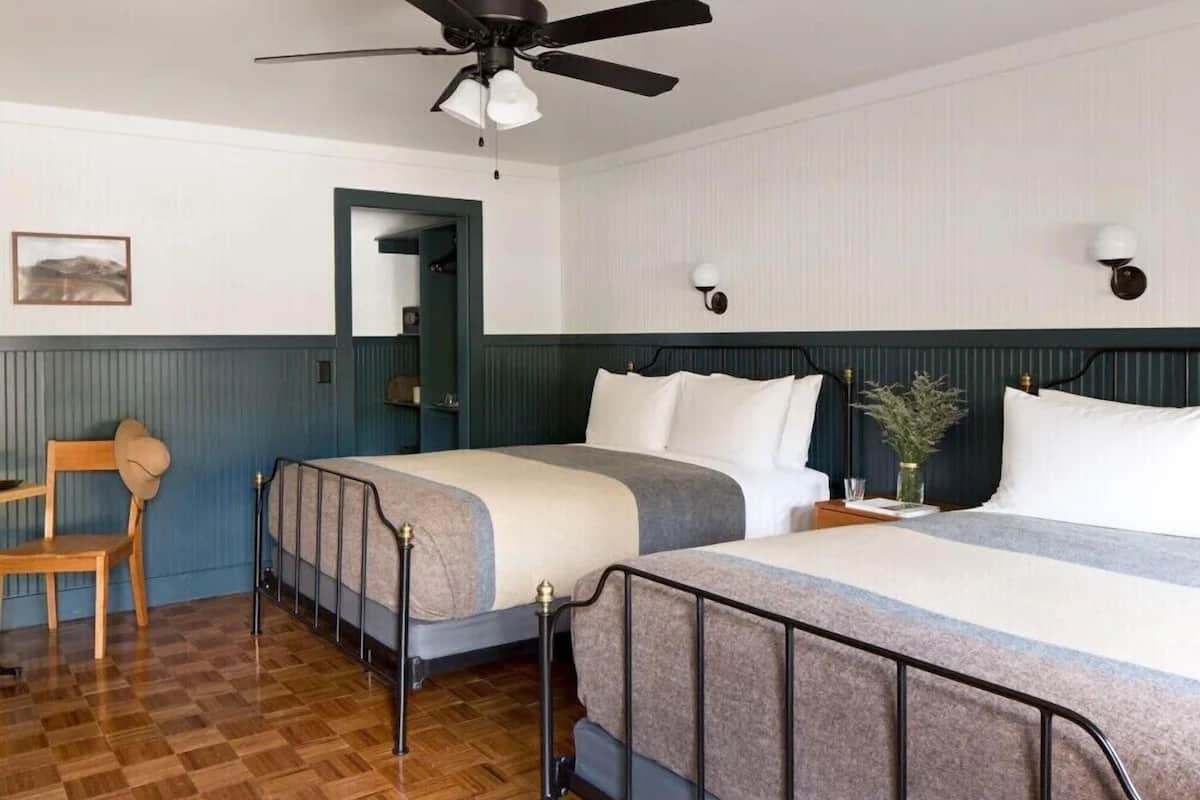 Une chambre avec deux lits, un ventilateur de plafond, une chaise en bois et une petite table sur laquelle se trouve un vase.