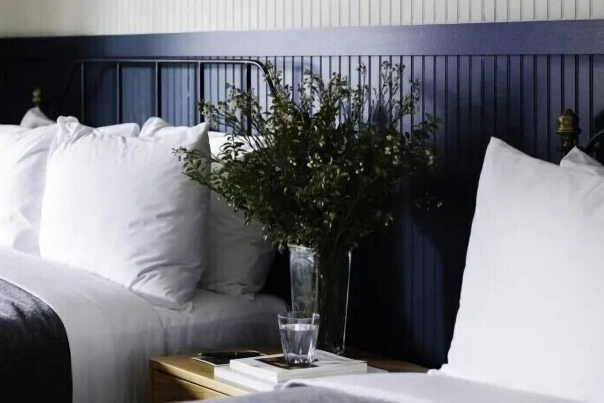 Un lit bien fait, avec du linge de lit blanc, un vase avec des plantes vertes et un verre d’eau sur une table de chevet.