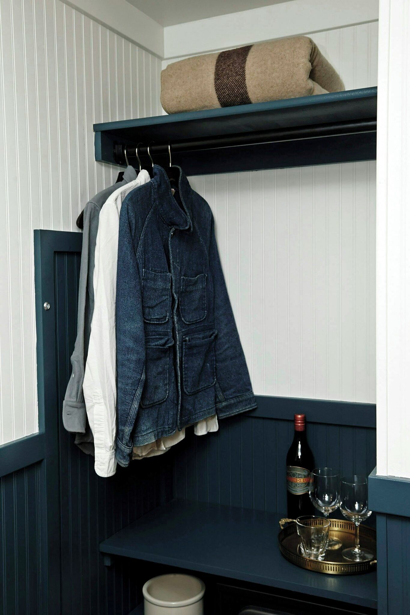 A room with a dark blue bench, a hanging coat rack with a denim jacket, a bottle of wine, and glasses on the bench.