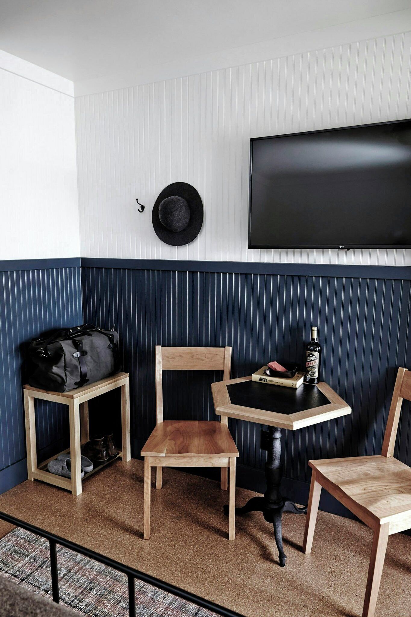 A room with a television mounted on the wall, a hat hanging on the wall, a small table with a bottle, and two chairs.