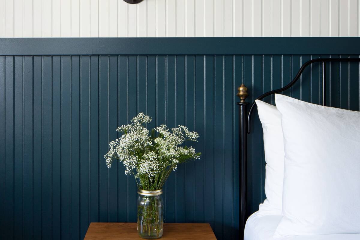 Un lit bien fait, avec du linge de lit blanc, une tête de lit bleu foncé et une table de chevet ornée d’un vase de fleurs.