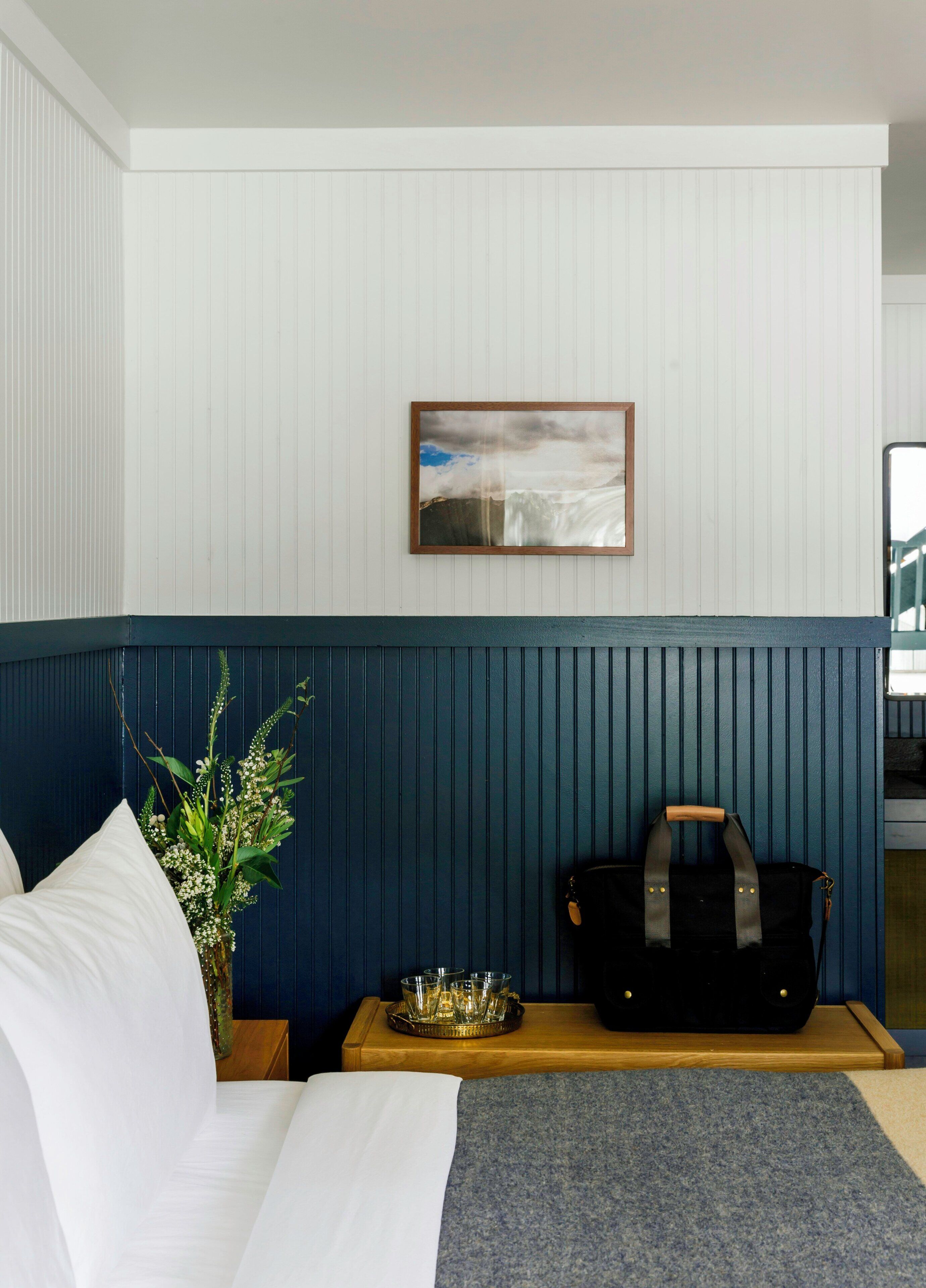 Une chambre d’hôtel avec un lit, une table de chevet ornée d’un vase de fleurs, un plateau avec des verres et un sac posé sur la table.