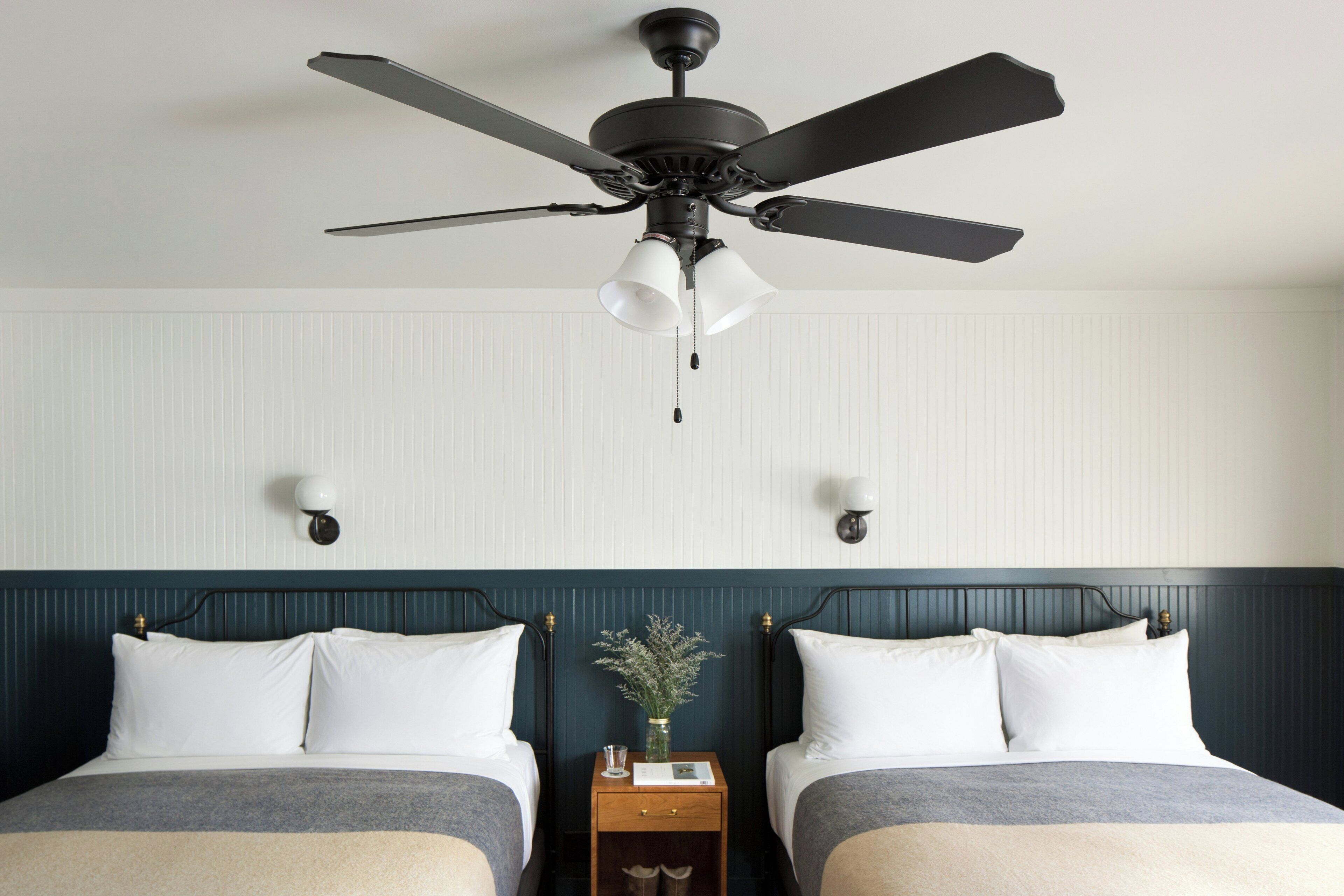 A hotel room with two beds, a ceiling fan, wall-mounted lights, and a nightstand with a vase.