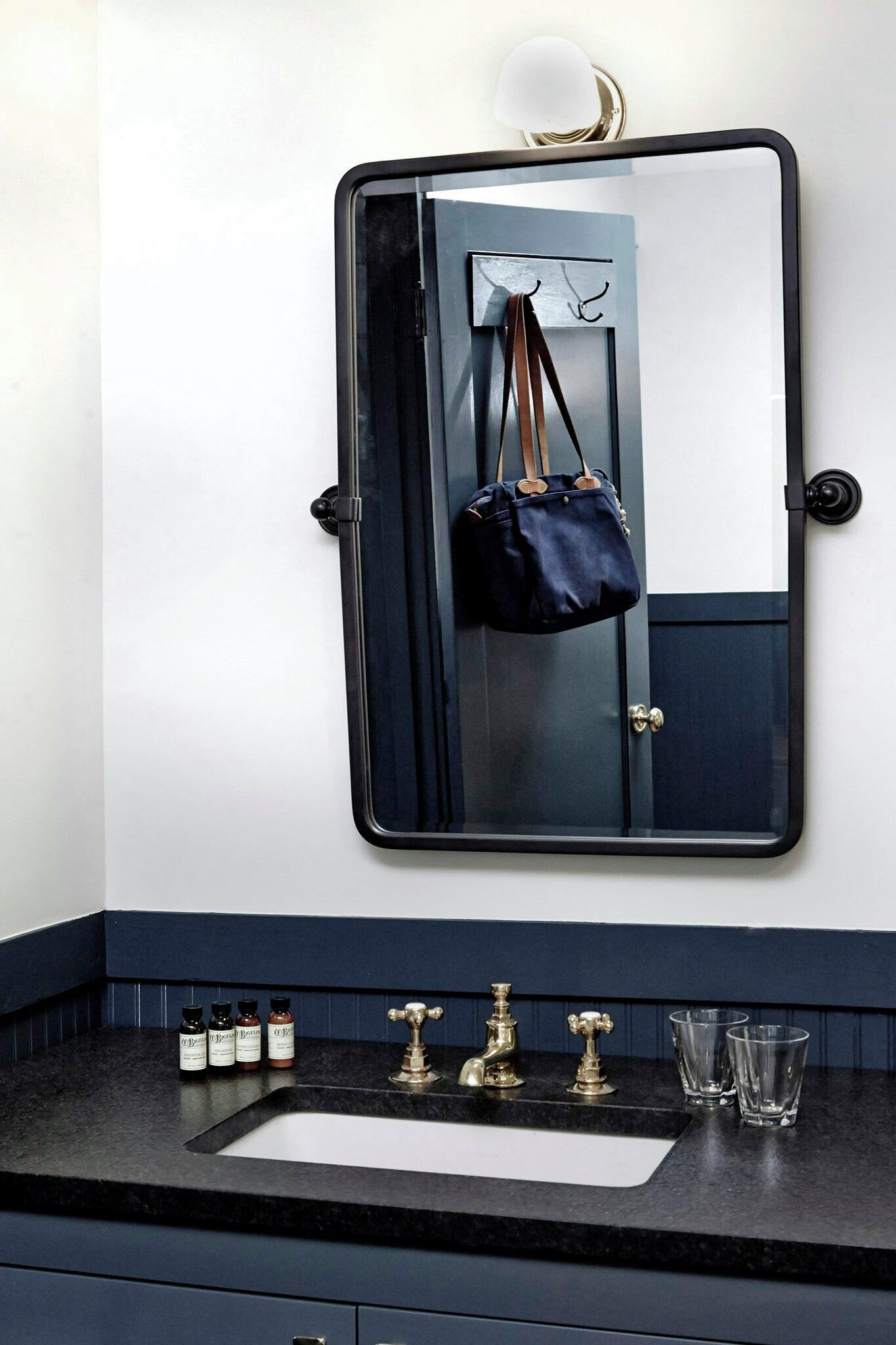 A bathroom with a dark blue vanity, a mirror with a black frame, a hanging bag, a sink with two faucets, and a bottle of hand soap.