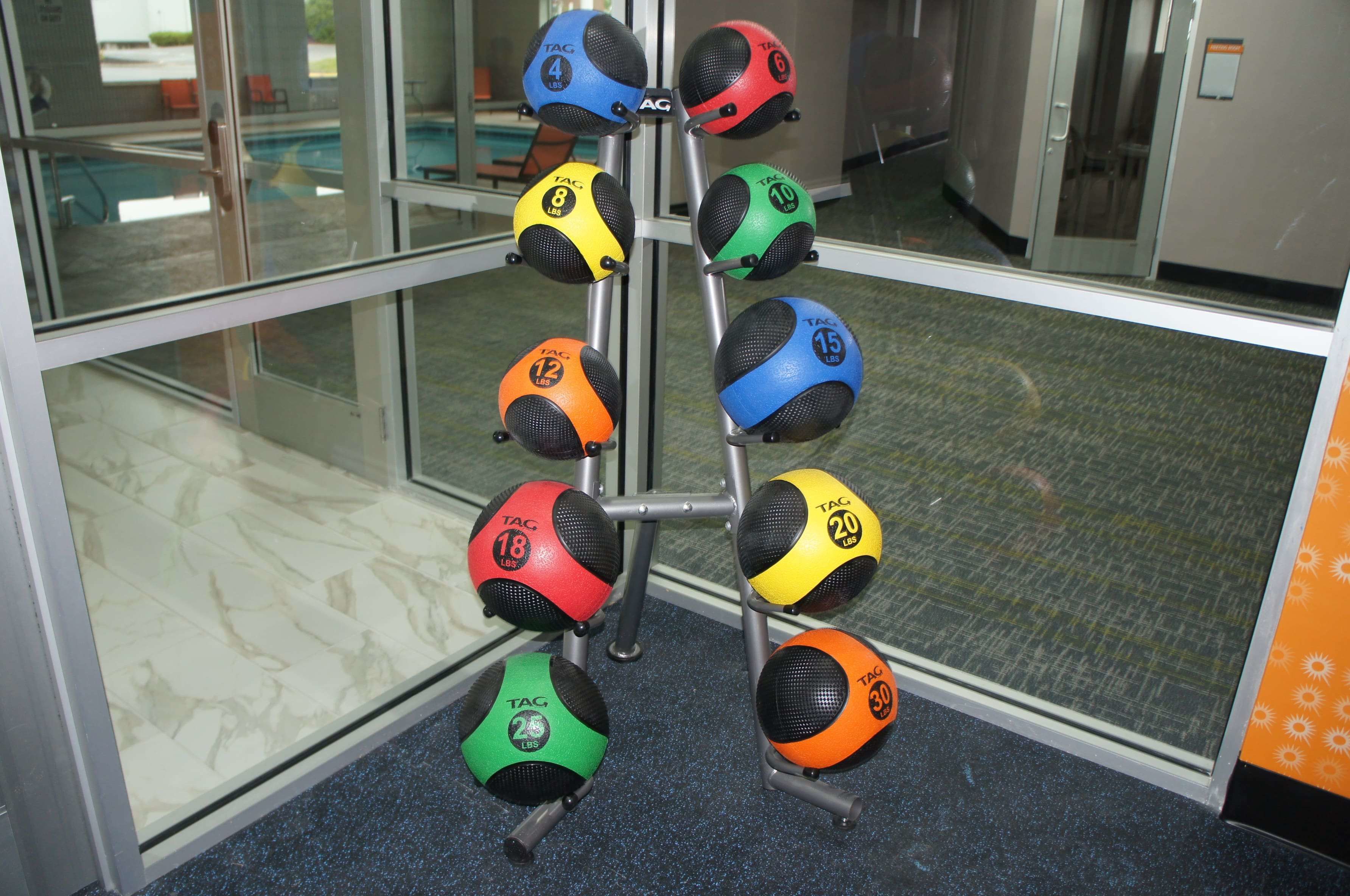 Una estructura con pelotas de ejercicio de diferentes pesos y colores, ubicada en una habitación con pared de vidrio y piso de baldosas.