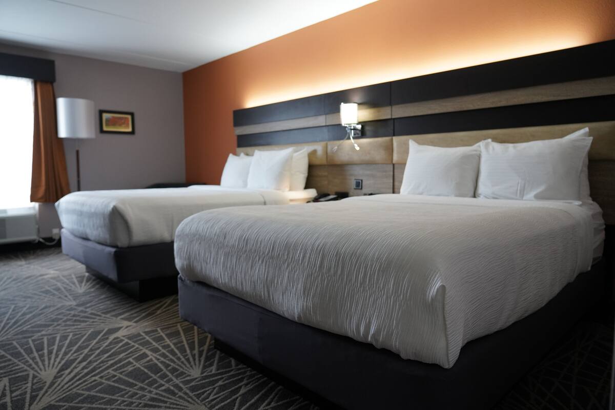 A hotel room with two beds, a headboard with a light fixture, a framed picture on the wall, and a patterned carpet.