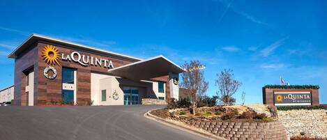 A La Quinta hotel with a modern design, featuring a prominent entrance and a landscaped area with a stone wall and plants.