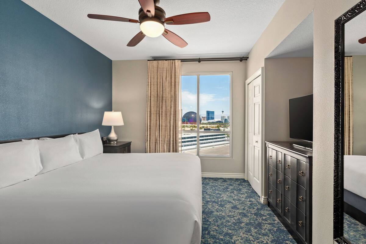 Habitación de hotel con una cama grande, ventilador de techo, televisor, cómoda y ventana con vista a la ciudad.