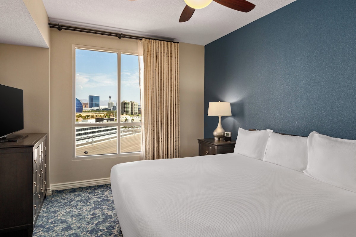Habitación de hotel con una cama grande, un televisor en una cómoda y una ventana con cortinas, con vistas a edificios.