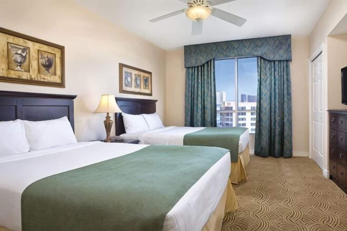 Habitación de hotel con dos camas, ventilador de techo, ventana con cortinas y vistas a la ciudad.