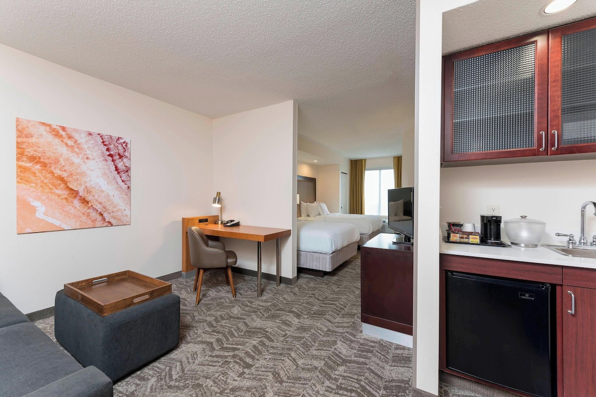 A hotel room with a bed, a desk, a chair, a small kitchenette, and a painting on the wall.