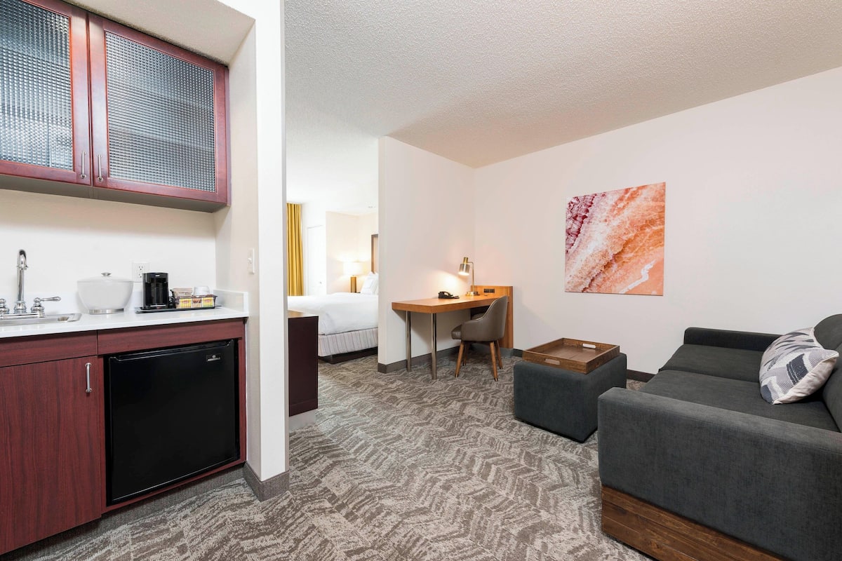 A hotel room with a kitchenette, a bed, a desk, a couch, and a painting on the wall.