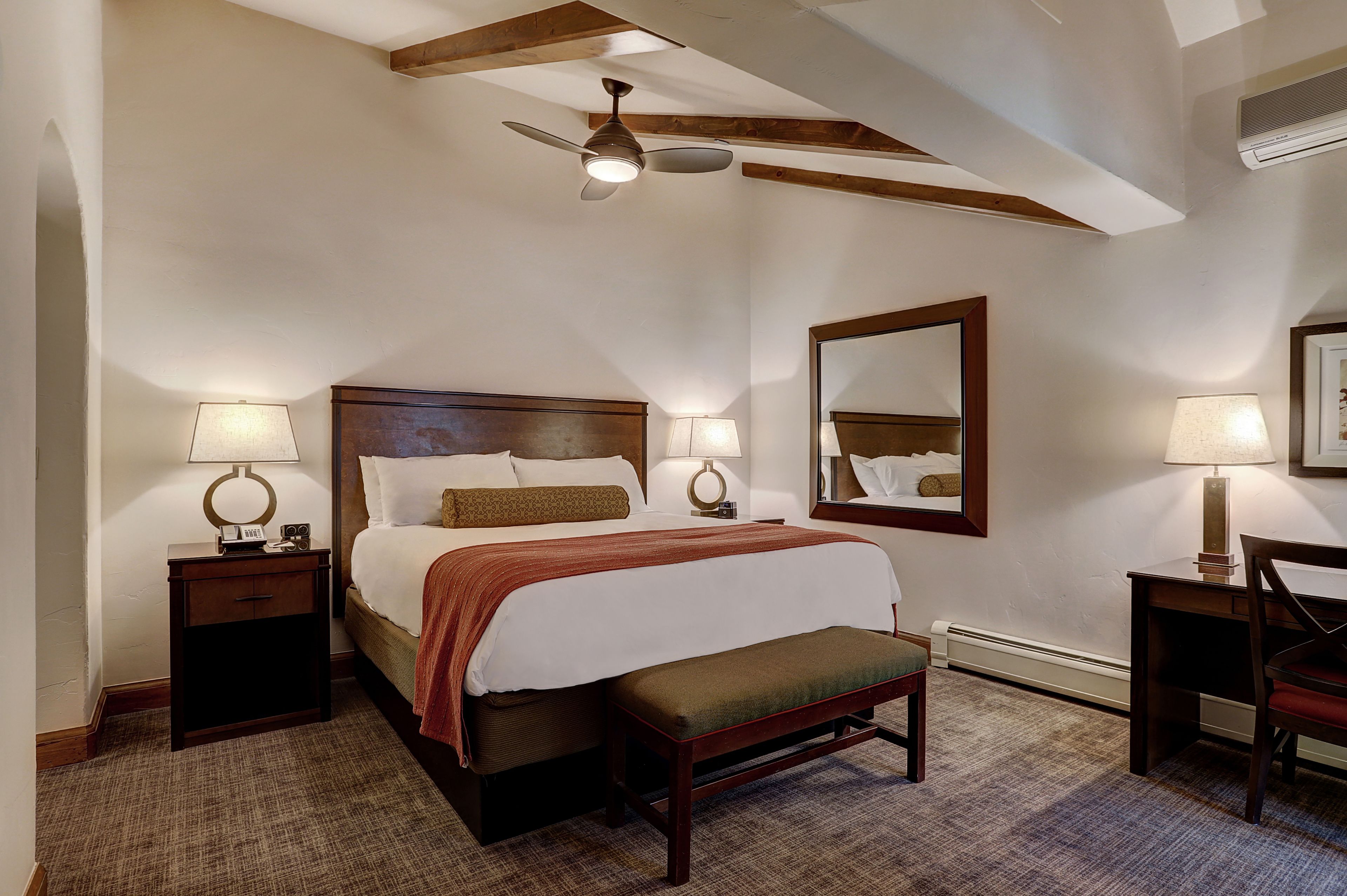 A hotel room with a large bed, two bedside tables with lamps, a bench, a ceiling fan, and a large mirror on the wall.