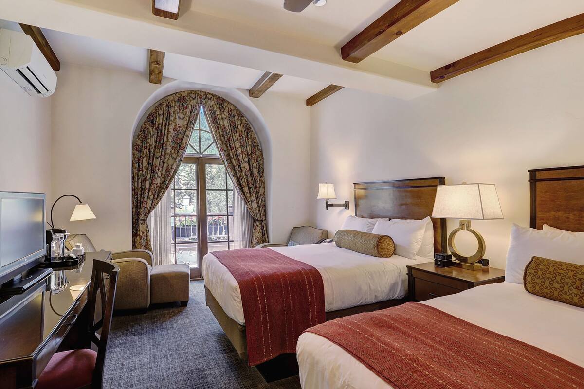 A hotel room with two beds, a desk with a chair, a TV, and a window with curtains.