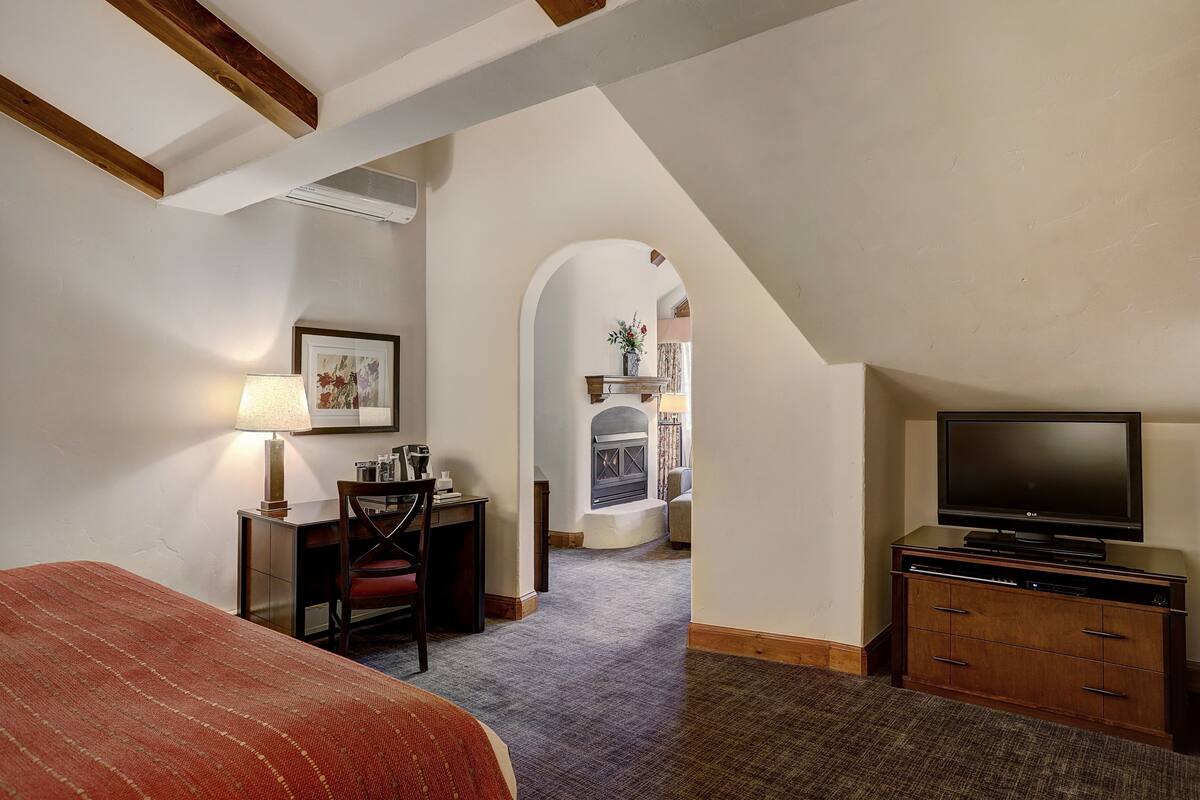 A hotel room with a bed, desk, chair, TV, fireplace, and a hallway leading to another room.