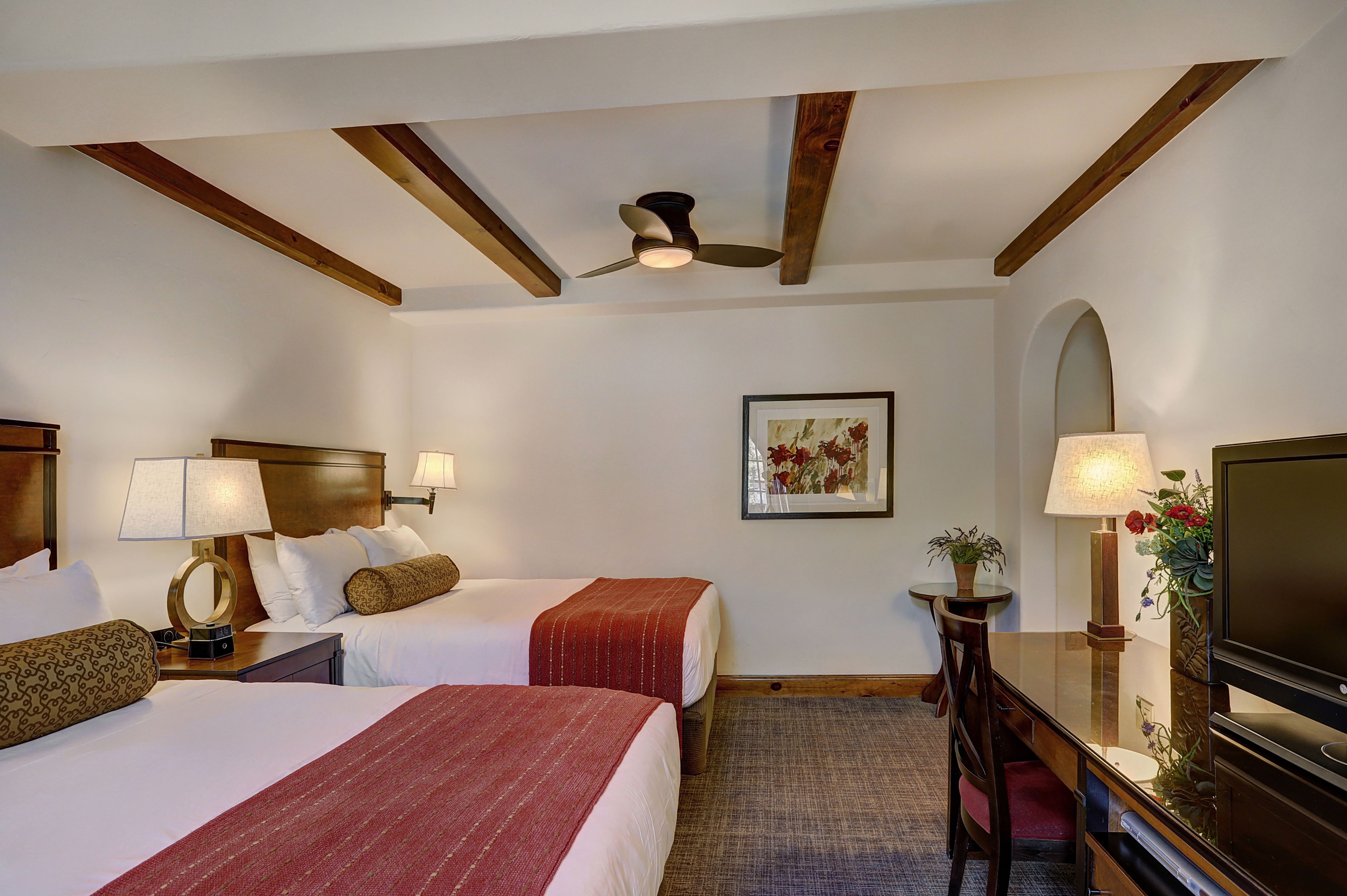 Quarto de hotel com duas camas, um ventilador de teto, um quadro, uma televisão e uma escrivaninha com abajur.