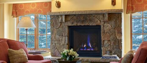 A cozy living room with a stone fireplace, red upholstered chairs, and a view of snow-covered trees through large windows.
