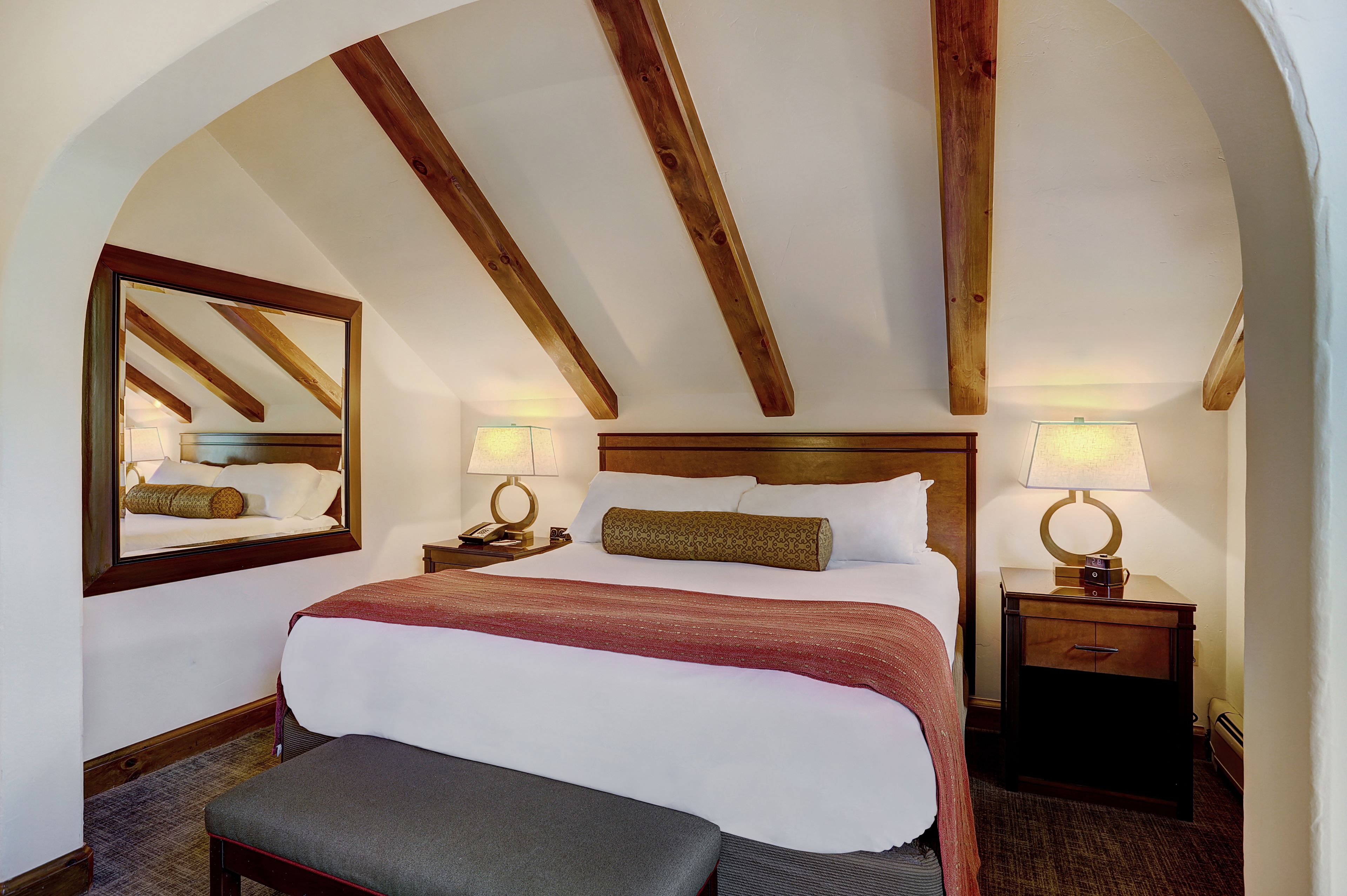 A hotel room with a large bed, wooden beams on the ceiling, a bedside table with a lamp, and a bench.
