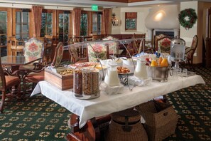 A buffet setup with various food items and beverages on a table, surrounded by wooden chairs and tables in a room with patterned carpet and large windows. - Austria Haus (Vail)