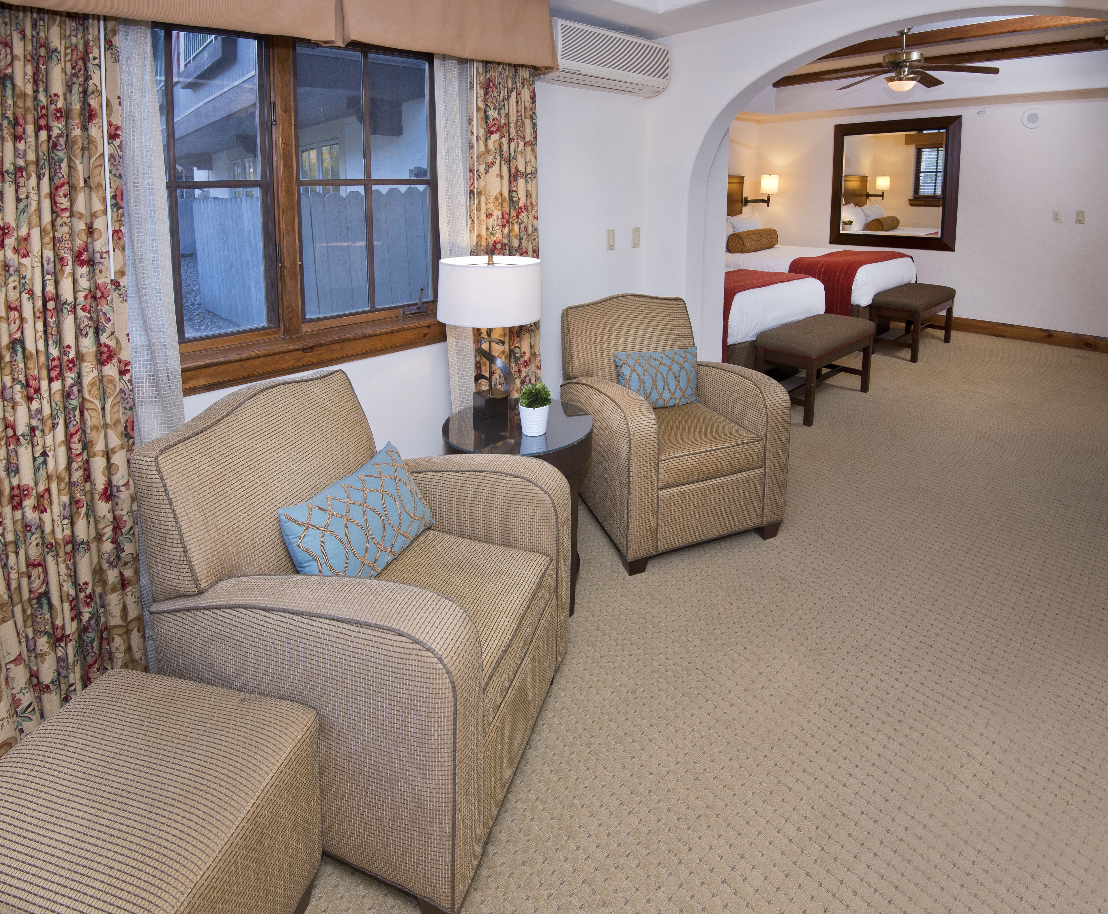 A hotel room with a sofa, armchair, and a small table with a lamp. There is a window with curtains, a ceiling fan, and a mirror on the wall.