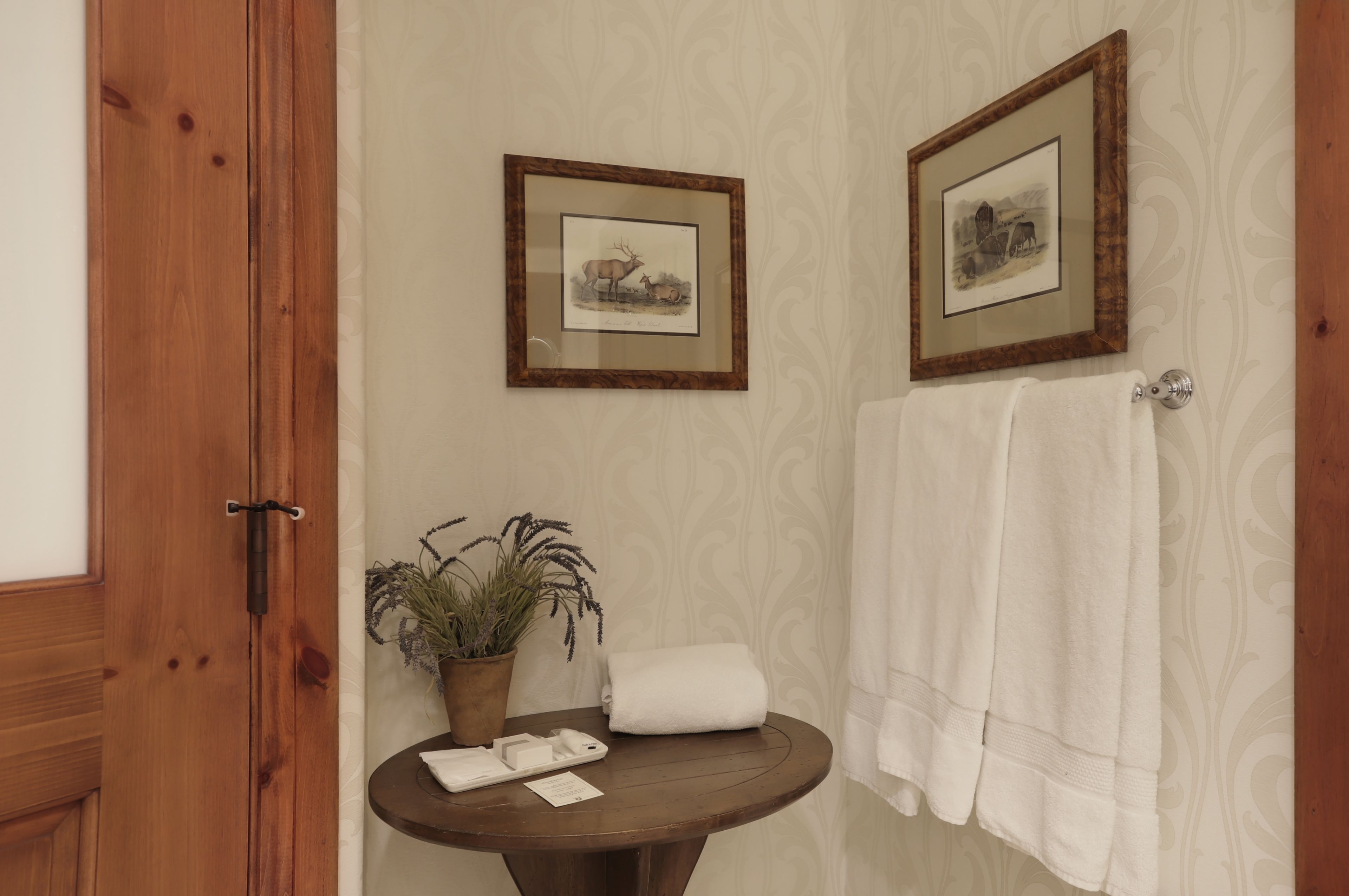 A bathroom with wooden doors, a round wooden table with a potted plant, two framed pictures on the wall, and a white towel hanging on a towel rack.