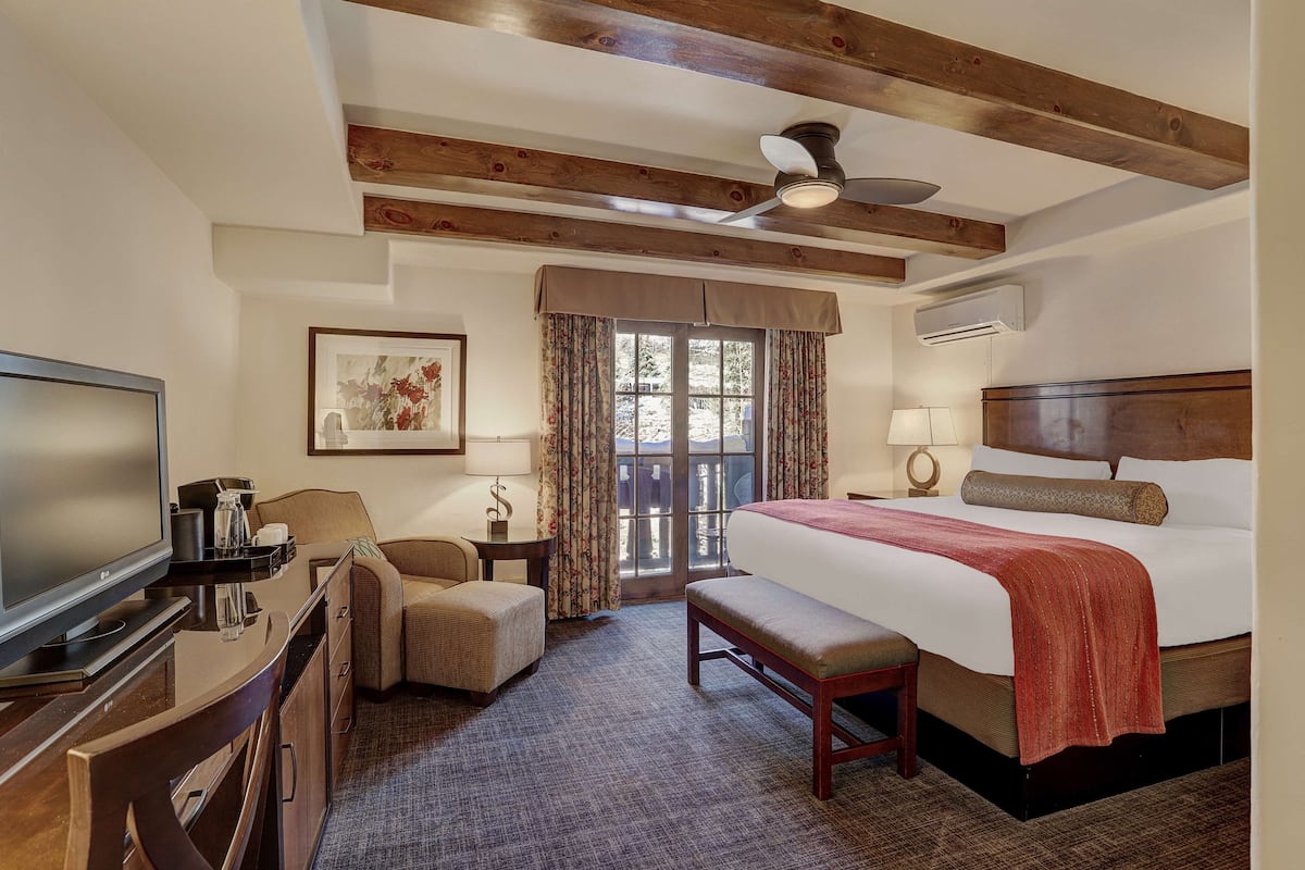 A hotel room with a large bed, a television, a desk with a computer, a chair, a bench, a window with curtains, and a ceiling fan.