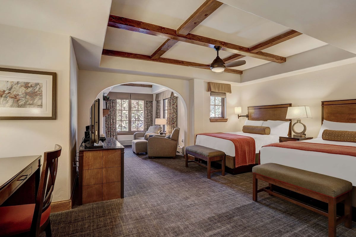 A hotel room with two beds, a desk, a chair, and a small table. There is a window with curtains, a ceiling fan, and a framed picture on the wall.