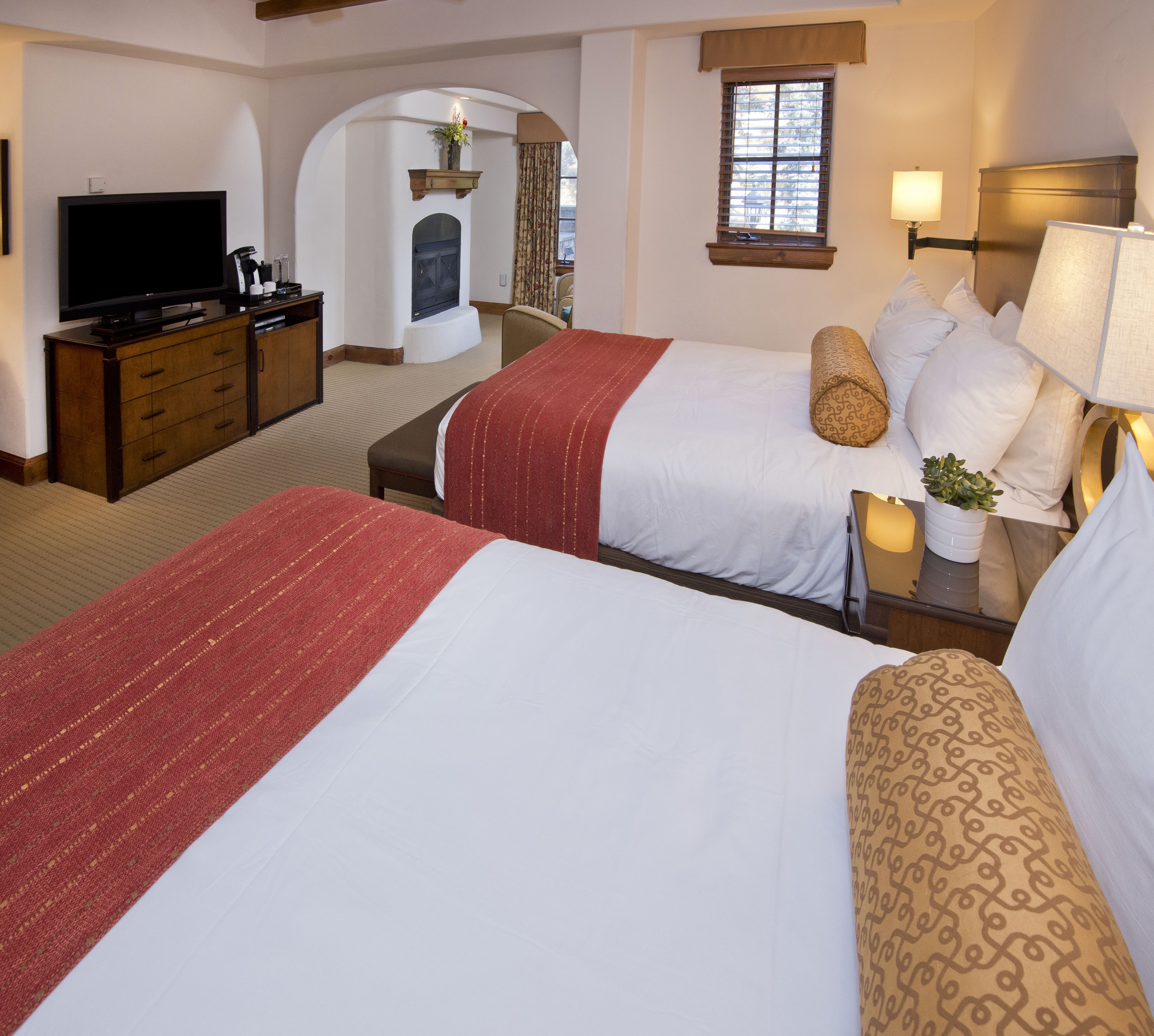 A hotel room with two beds, a TV, a fireplace, and a window with blinds.