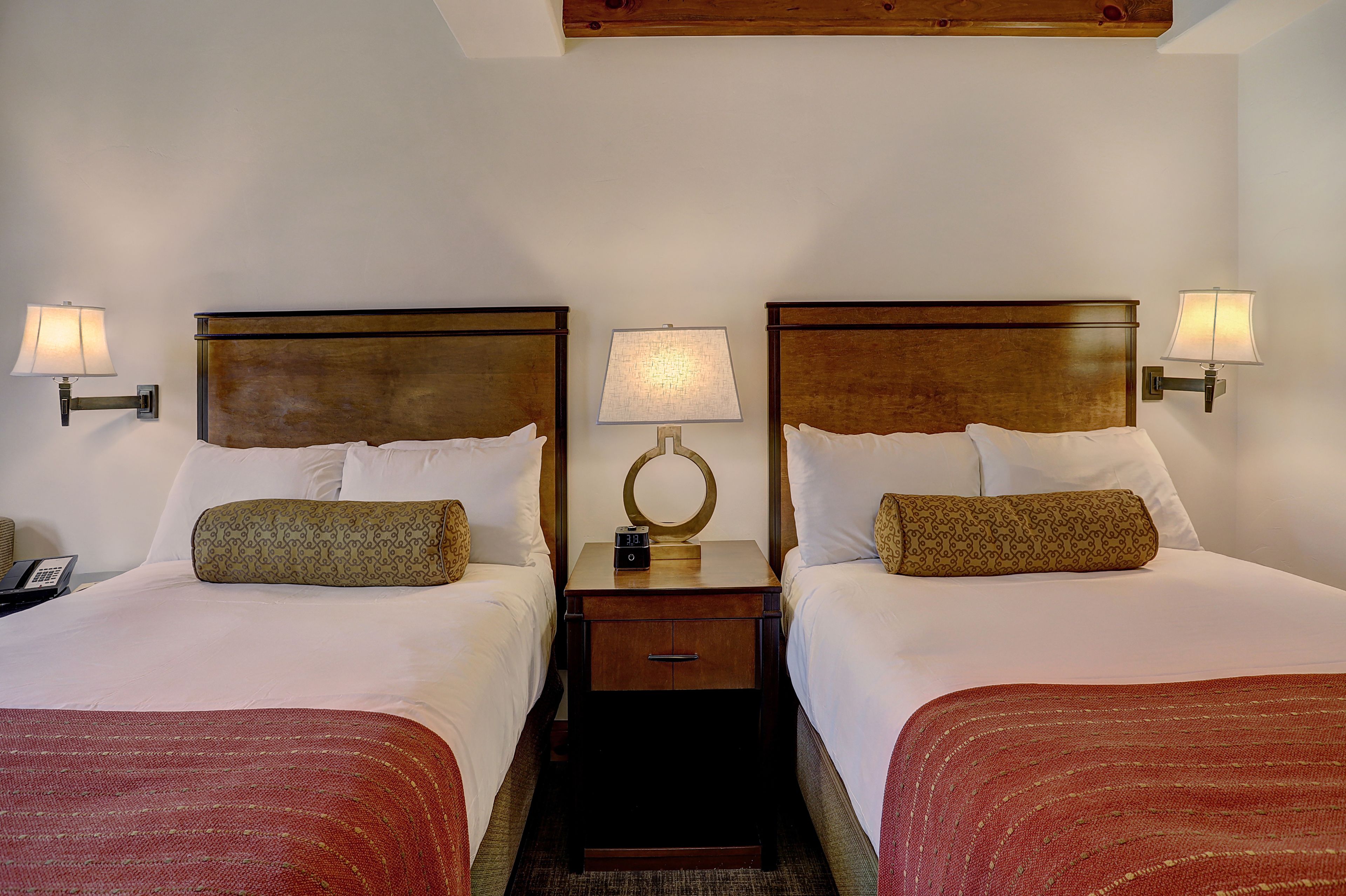 A hotel room with two beds, each with a wooden headboard, white linens, and a red throw blanket. There is a nightstand with a lamp between the beds, and a small table with a clock on it.