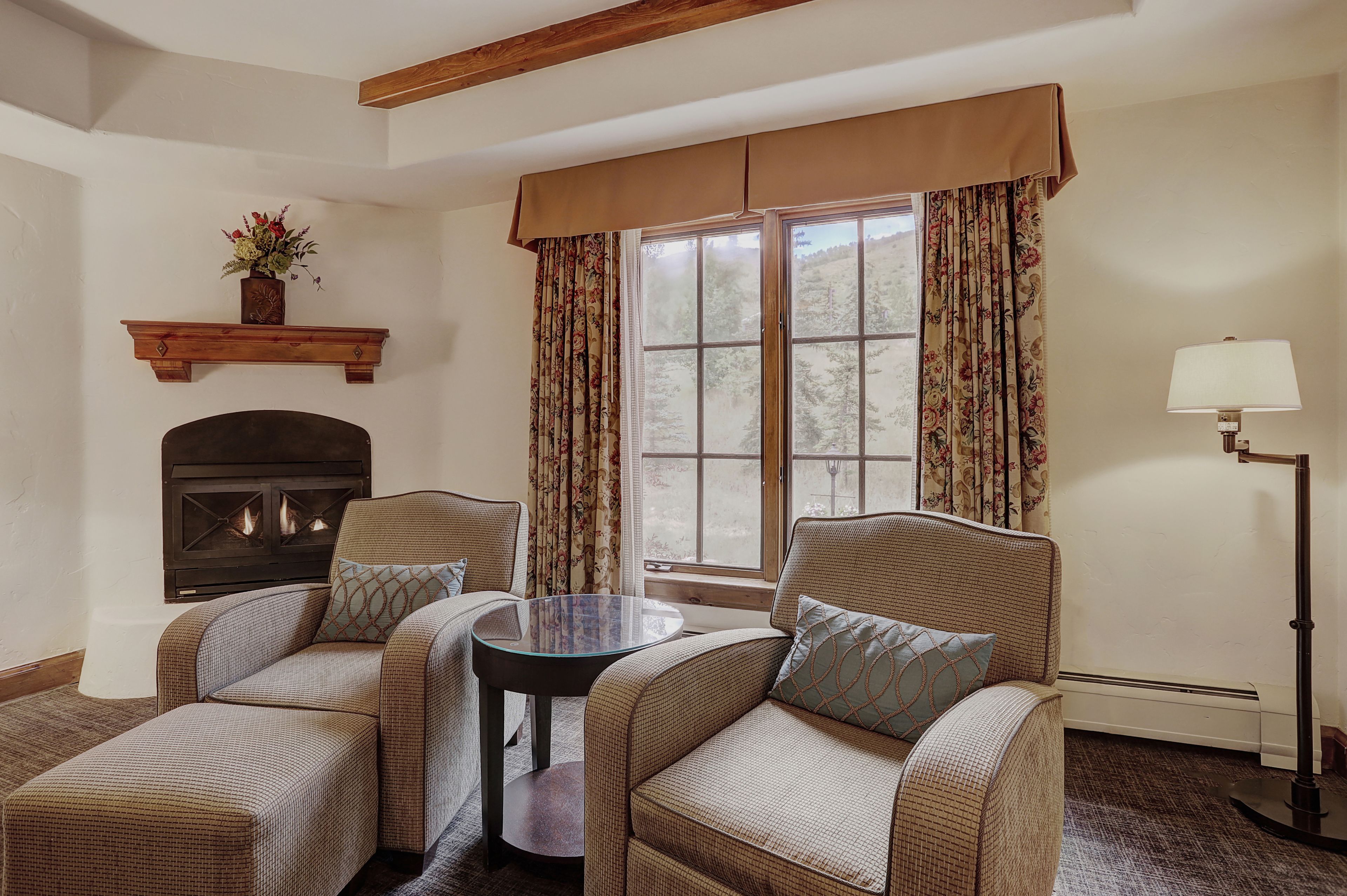 A cozy room with two armchairs, a small table, a fireplace, and a large window with curtains.