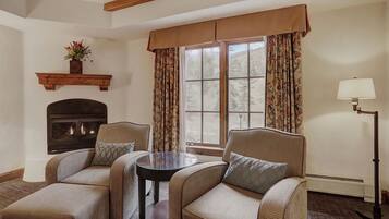A cozy room with two armchairs, a small table, a fireplace, and a large window with curtains.