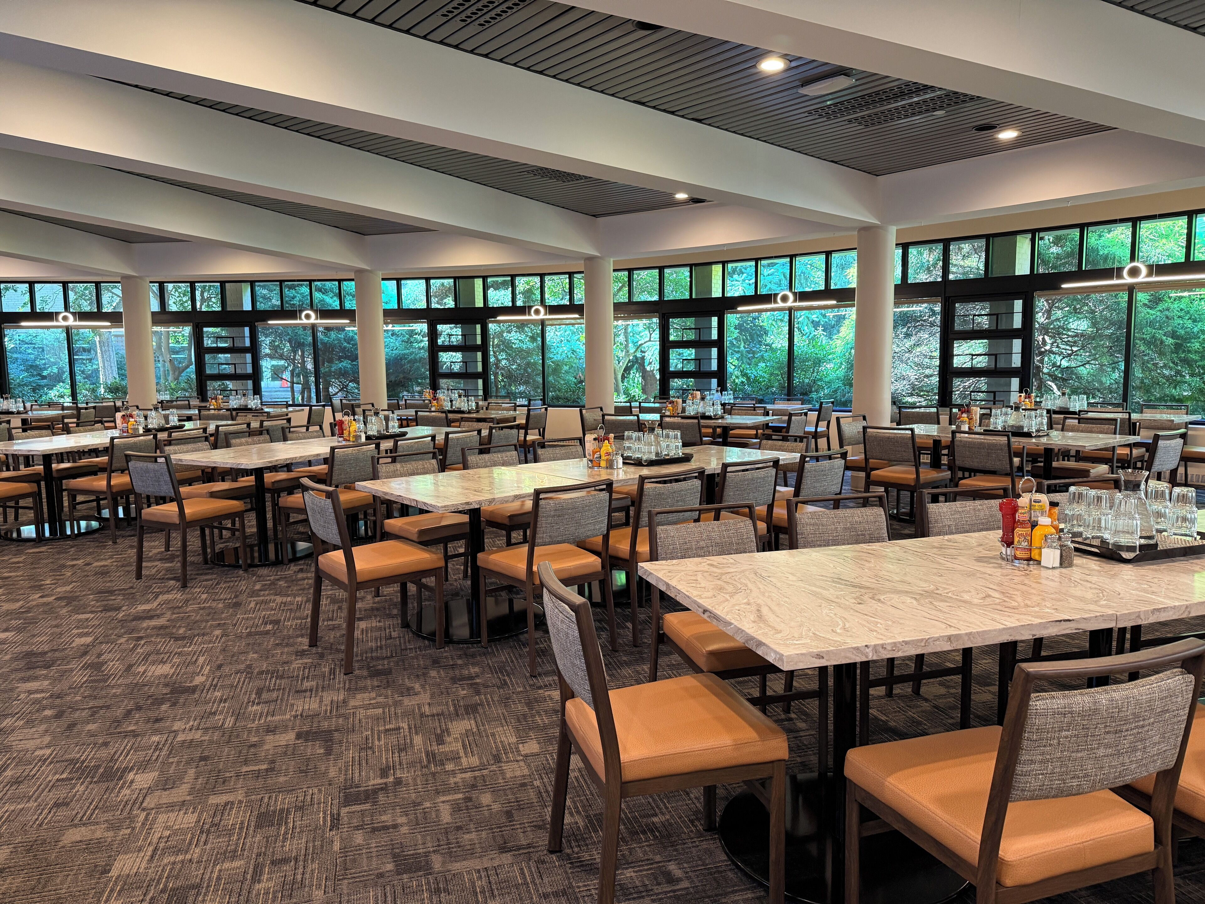 Un'ampia sala da pranzo con diversi tavoli e sedie, e grandi finestre che si affacciano sul verde circostante.