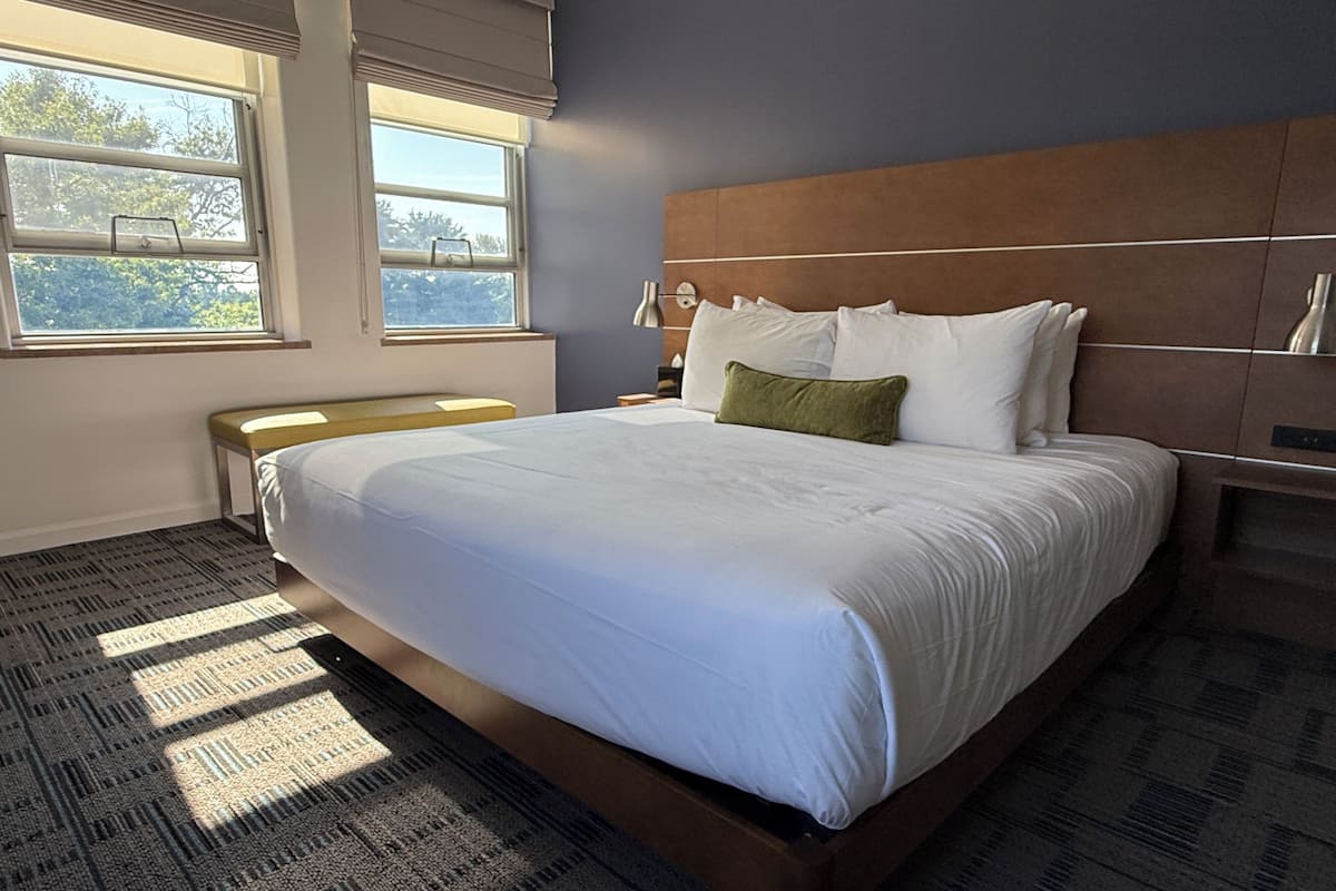 A large bed with a wooden headboard, a bench, and a window with blinds.