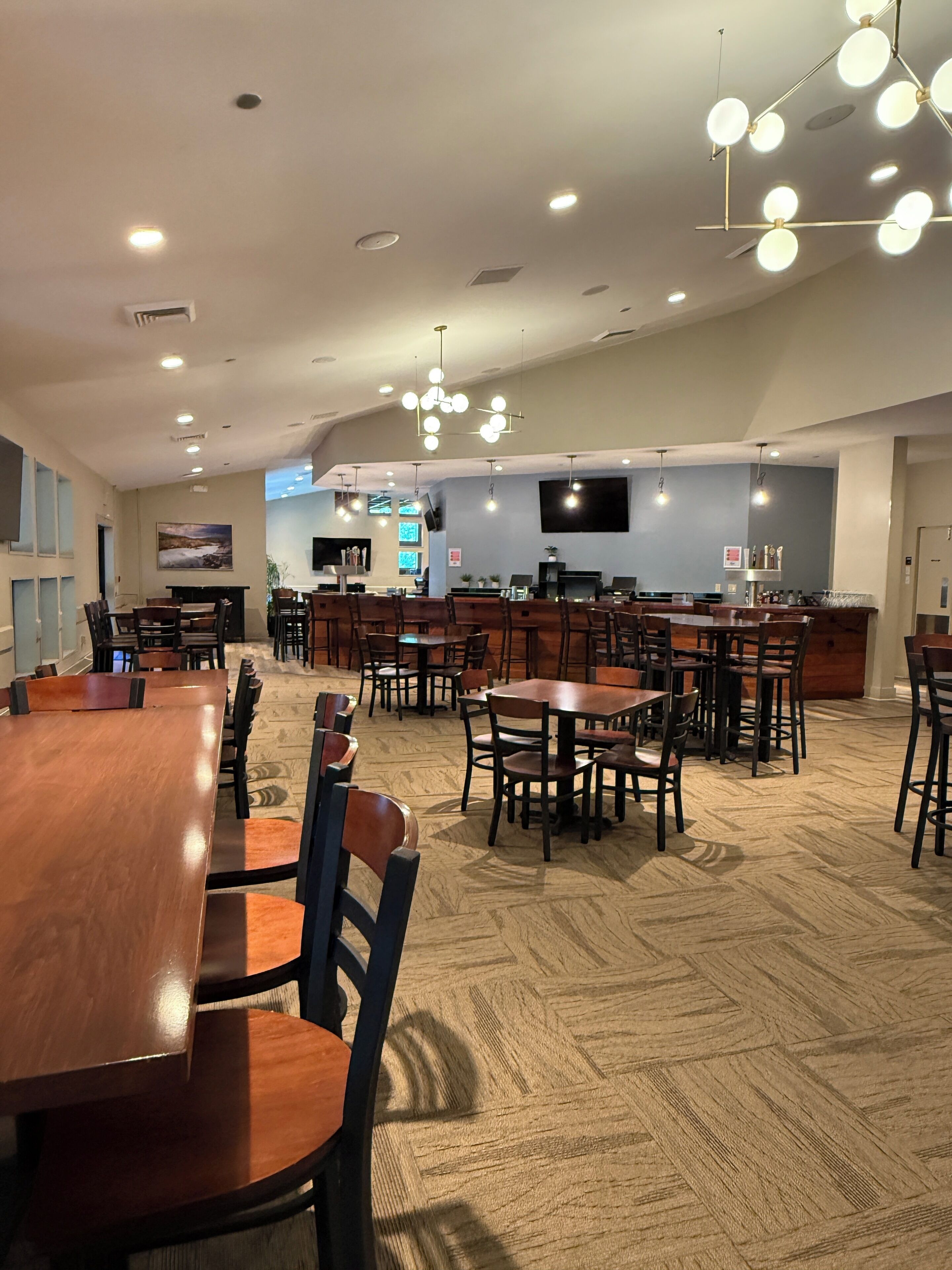 Un vaste espace dédié à la salle à manger, avec de nombreuses tables et chaises en bois, un bar situé au fond de la pièce et une moquette à motifs.